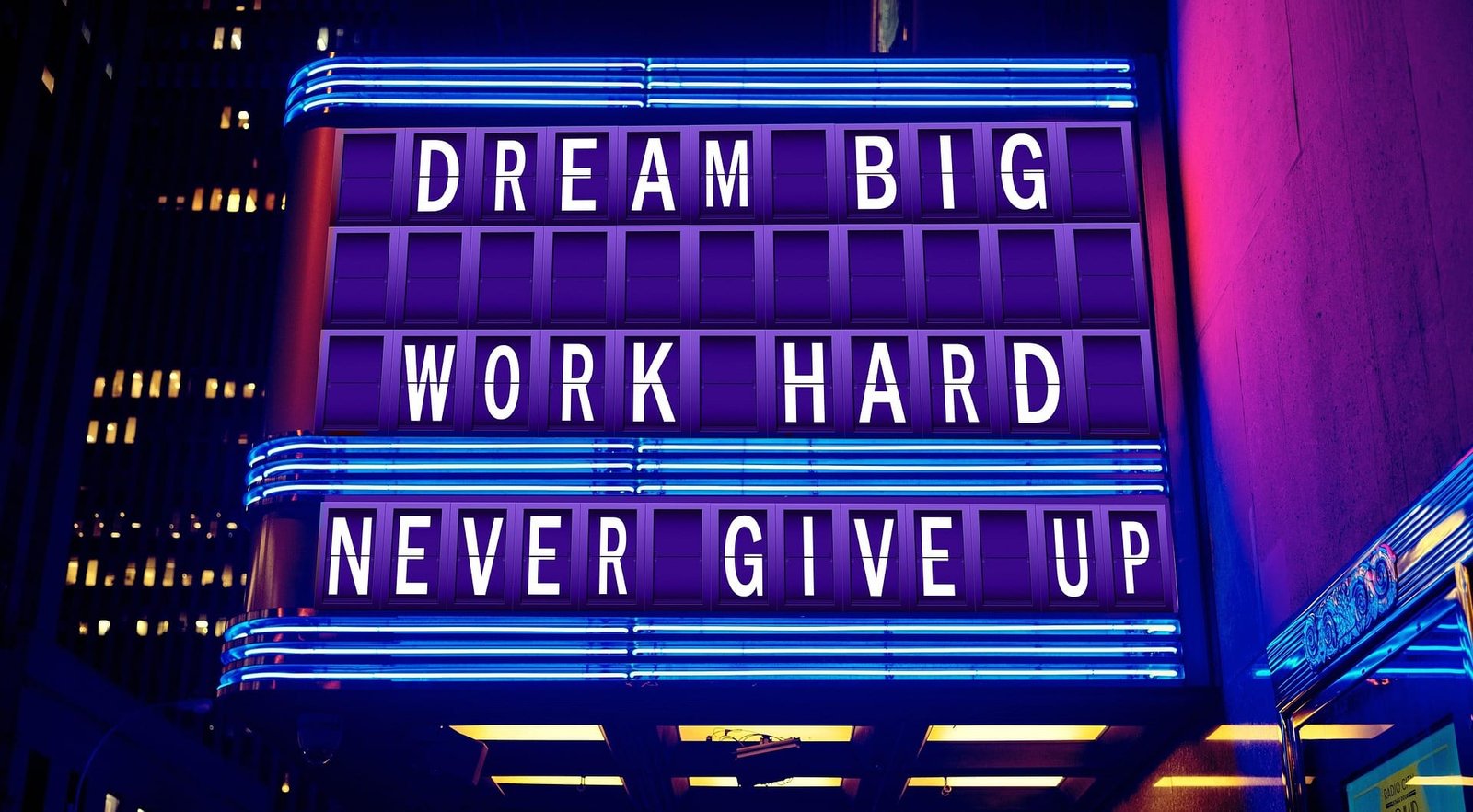 The image shows a neon signboard with a motivational message displayed in large, bold white letters against a purple background. The signboard is illuminated with blue neon lights and is set against a dark urban backdrop with some building lights visible in the background. The message on the signboard reads: DREAM BIG
WORK HARD
NEVER GIVE UP